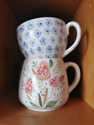 Eine blaue Tasse mit Blumenmuster steht auf einer weißen Tasse mit Pilzmuster.